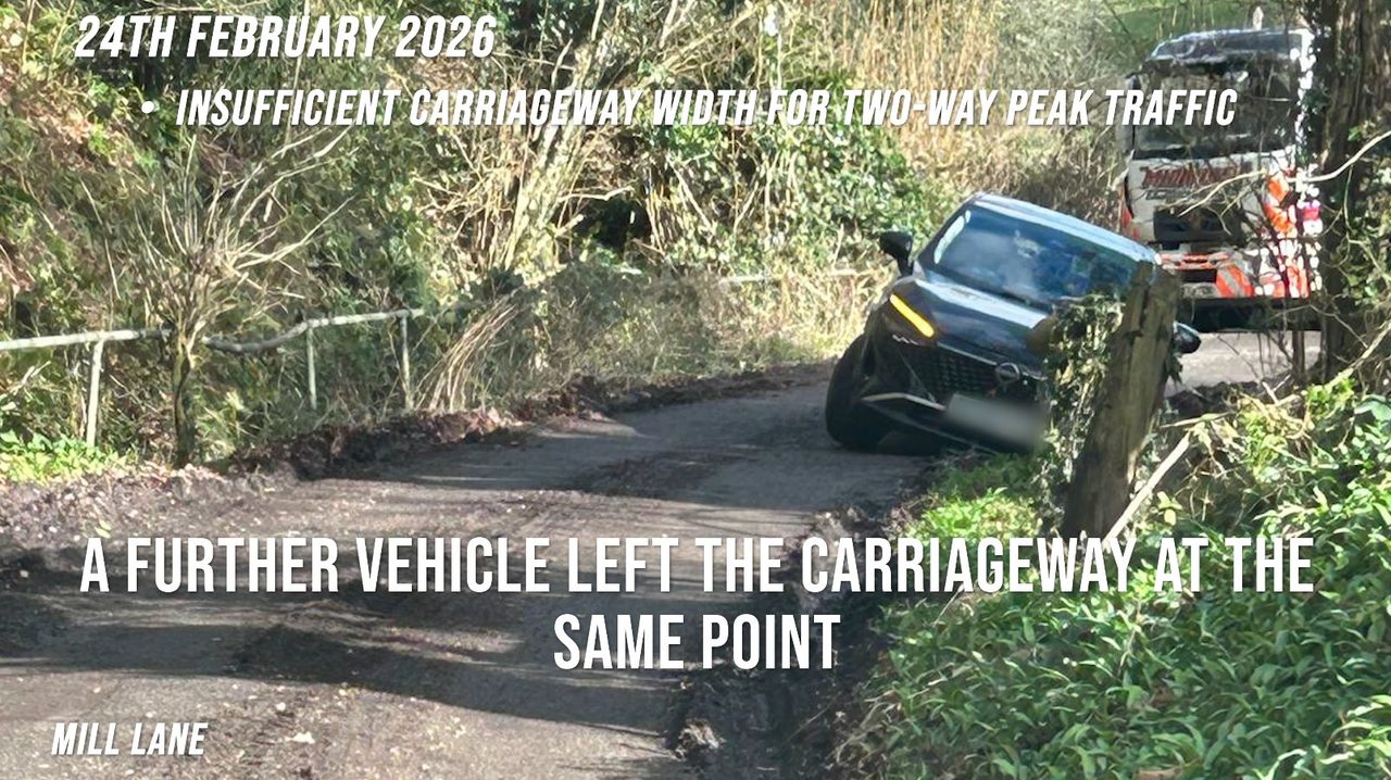 Mill Lane, 24 February 2026: a dark SUV with its front wheels off the carriageway against the verge, a recovery vehicle waiting behind on the narrow lane.